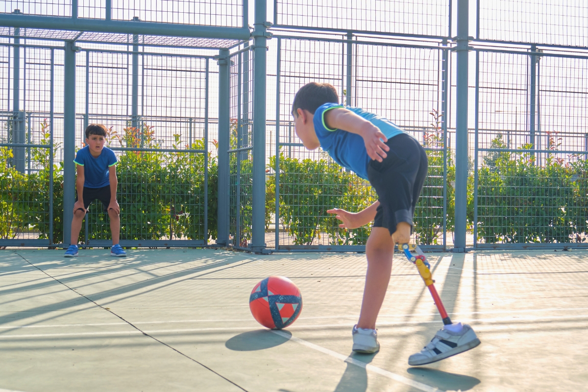 A young boy with a pediatric lower-limb prosthesis kicking a soccer ball on an outdoor court.