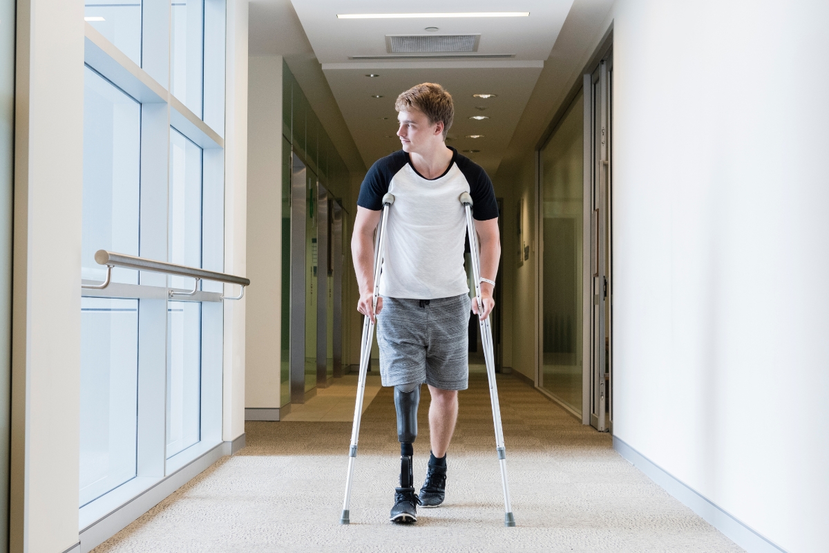 A teenage boy walking down a bright hallway with crutches and a lower-limb prosthesis.