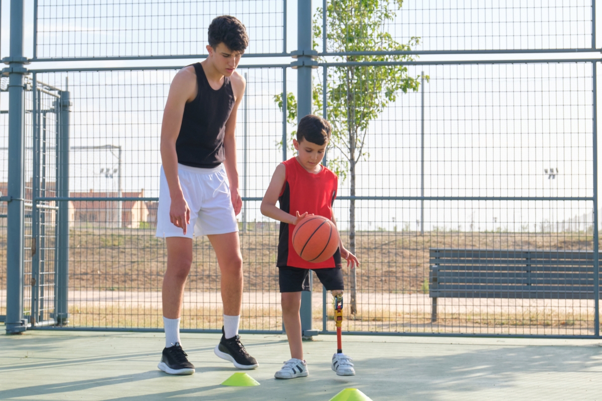 A young boy practicing basketball drills with an above-knee prosthesis on an outdoor court.