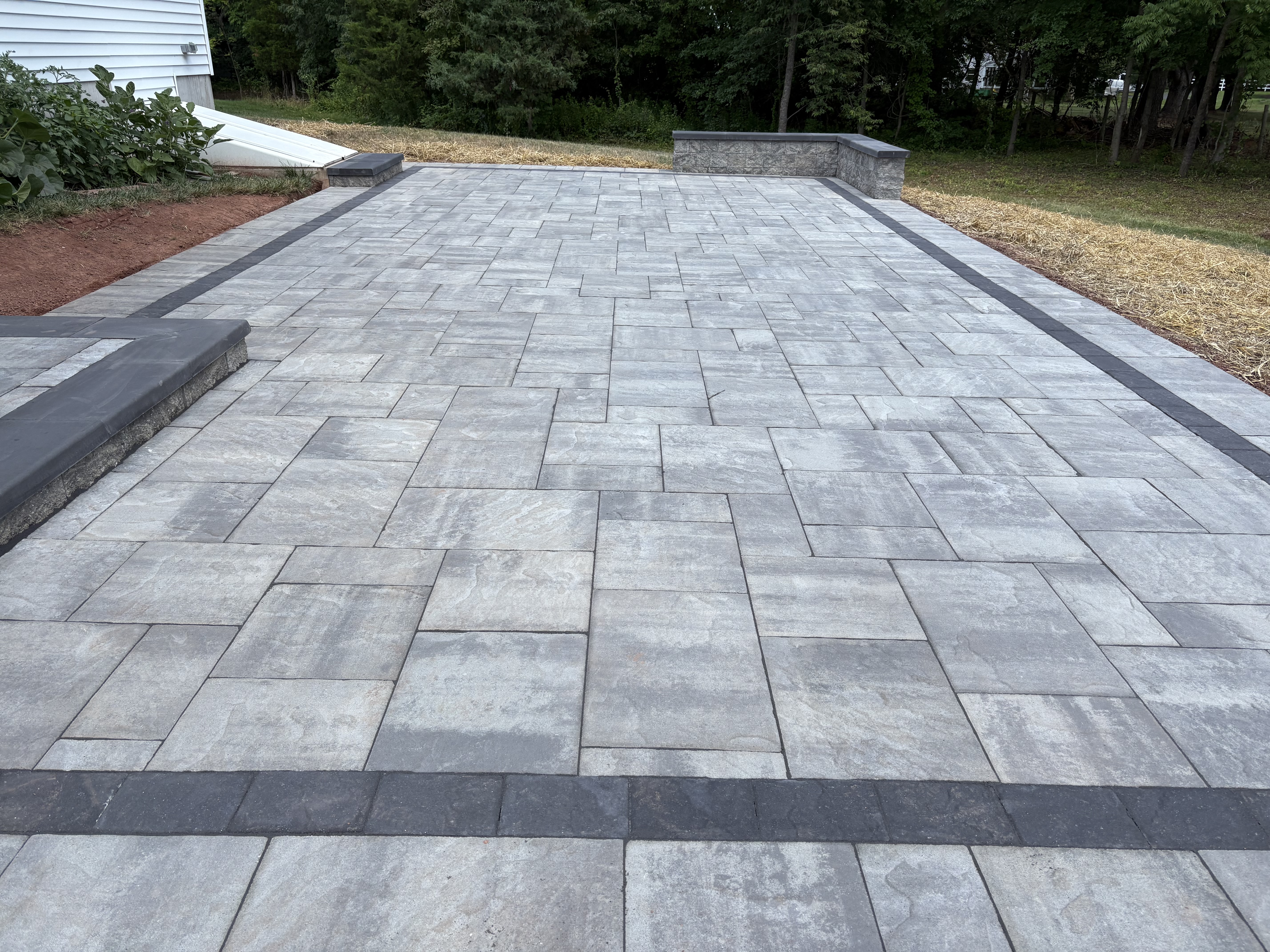 A Chester Springs, PA backyard patio showing reset and releveled concrete pavers with freshly repointed joints after hardscape repair. A stone retaining wall runs along the property edge with corrected grading visible at the base.