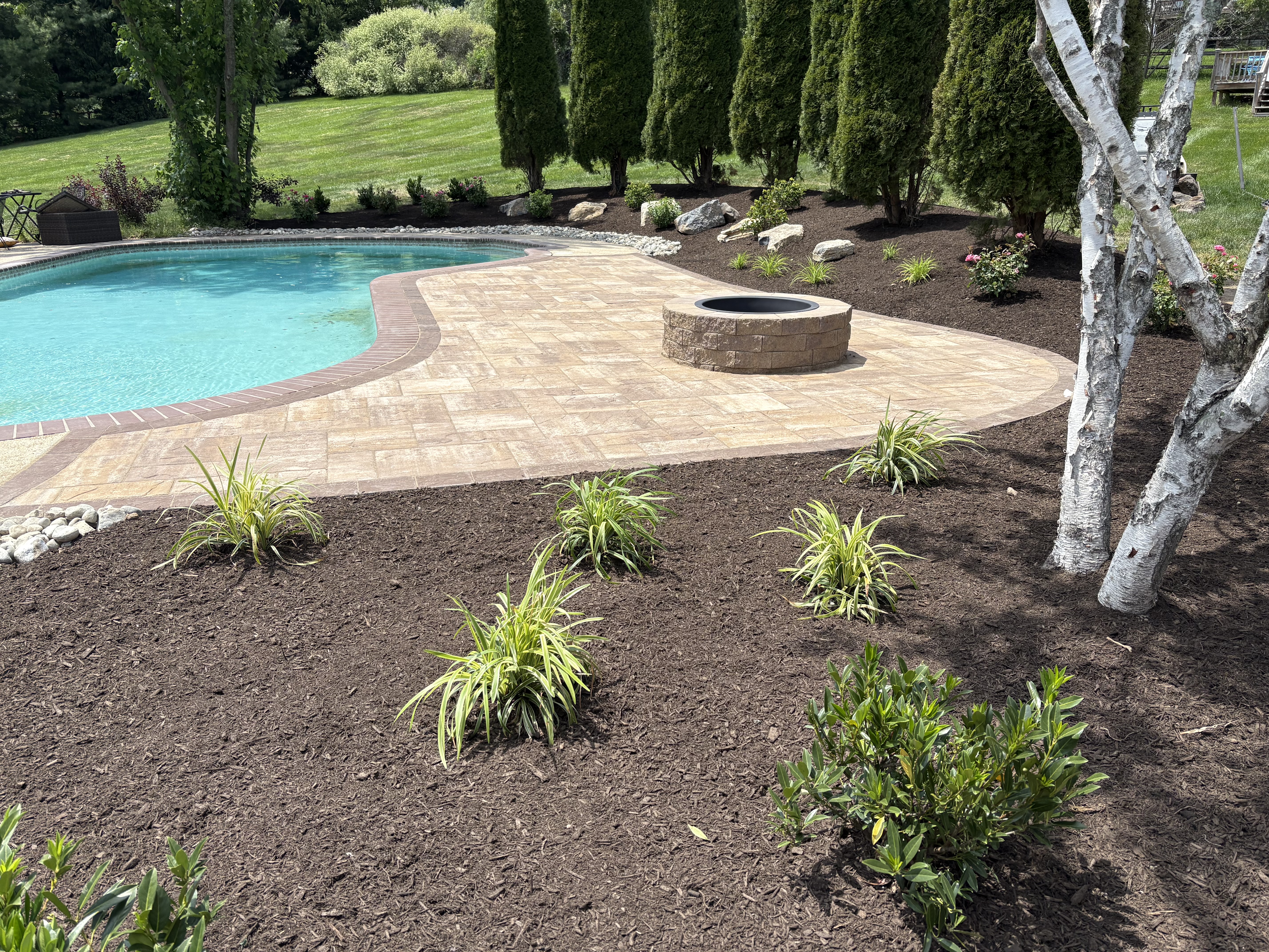 A finished stone patio installed by Fresh Look Landscape in a Chester Springs backyard, showing a level surface with clean edges set against a gently sloped, tree-lined yard. The patio includes an open seating area sized for outdoor dining and connects directly to the rear of the home.