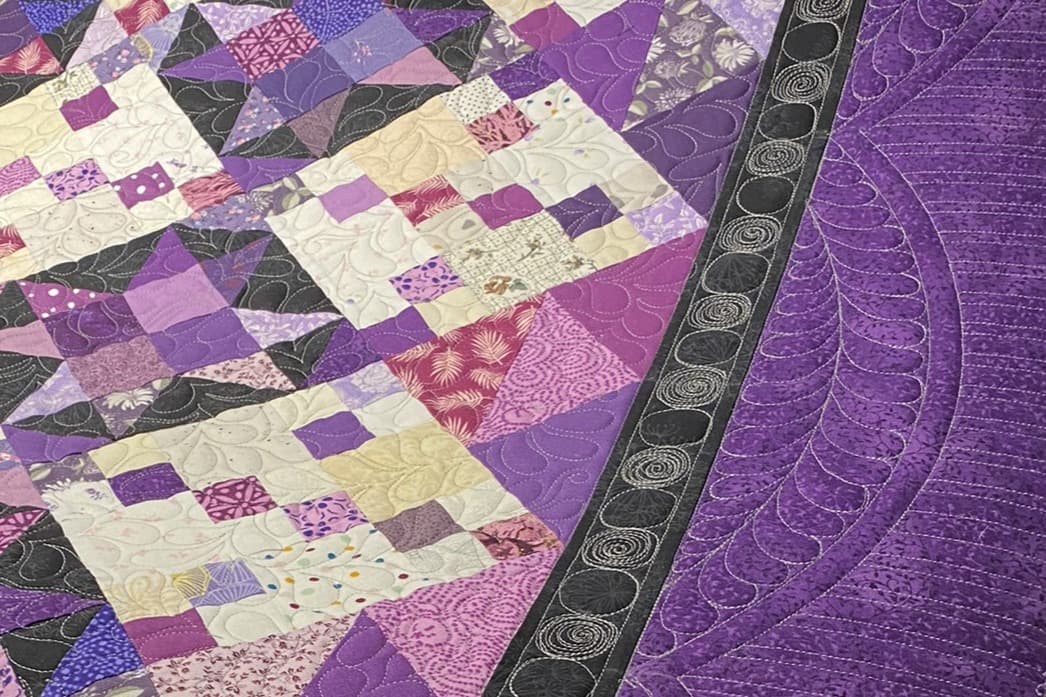 Detaljbilde av quiltingen på en lilla, svart og lys quilt. Det ses detaljert quilting i bordene og et heldekkende mønster i midtdelen