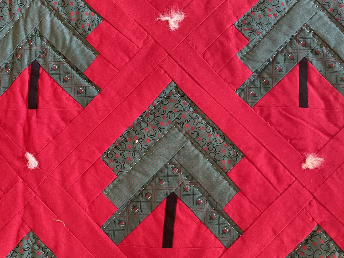 Utsnitt av en rød quilt med grønne juletrær i log cabin teknikk. Vi ser en hvit knute der blokkene møtes og håndquilting i juletrærne