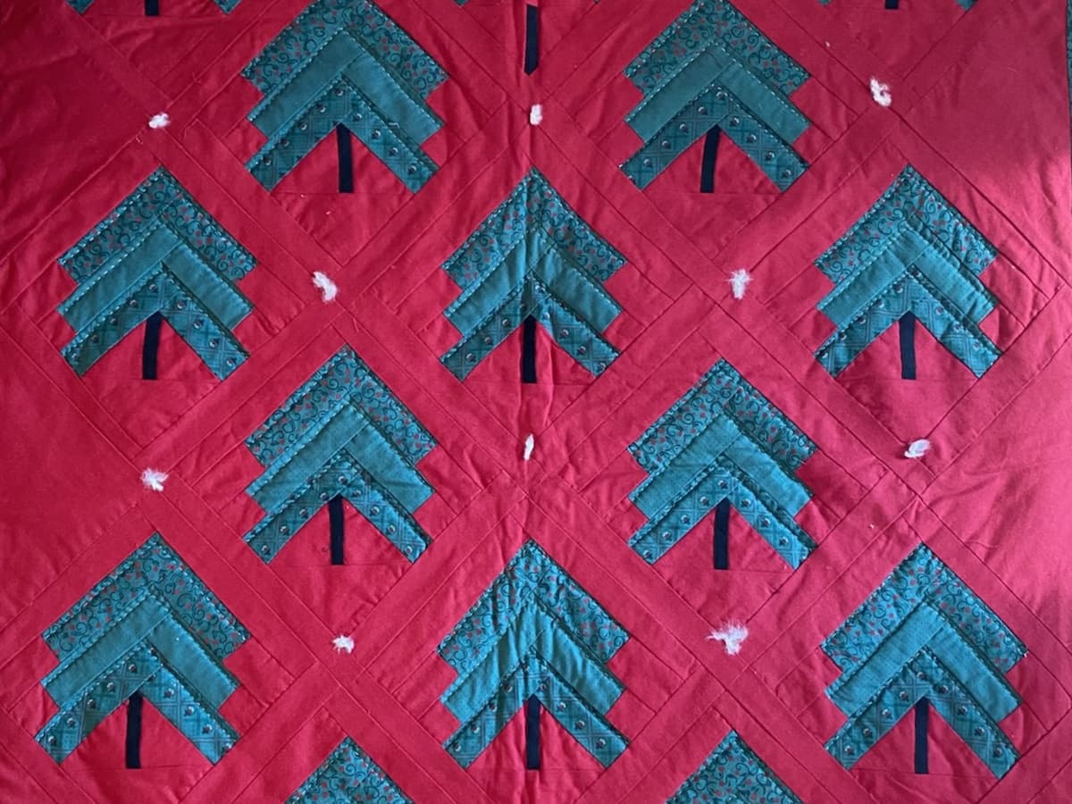 En quilt med grønne juletrær på rød bakgrunn. Quilten er satt sammen med hvite knuter der blokkene møtes