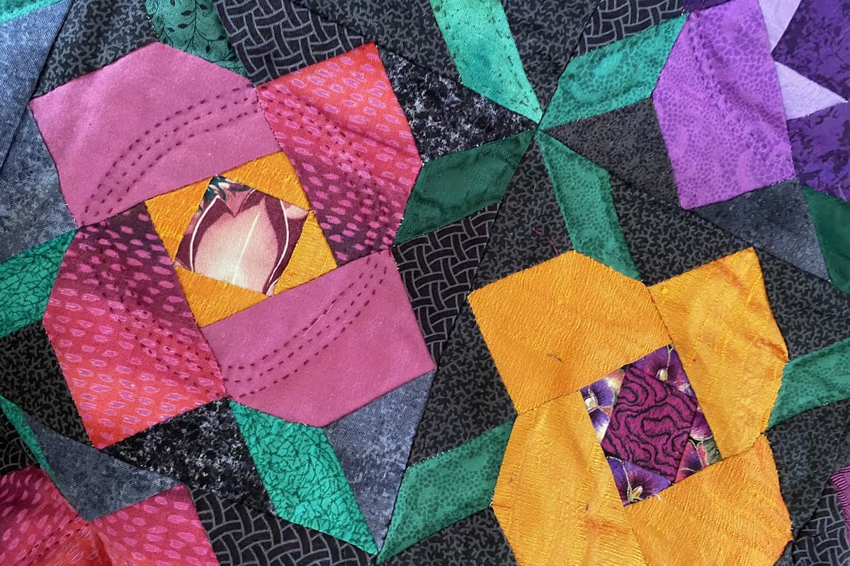 Nærbilde av en quilt med blokker som skal forestille blomster og blader. Mørk bakgrunn med rosa, gule og lilla fargermørk bakgrunn Nærbilde av en quilt med blokker som skal forestille blomster og blader. Mørk bakgrunn med rosa, gule og lilla fargermørk bakgrunn