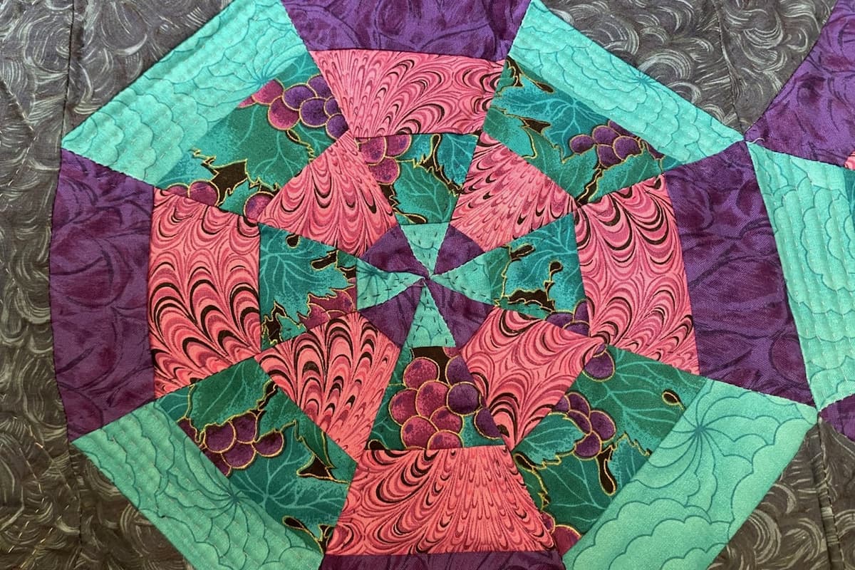 Nærbilde av en quilt fra 1980-tallet i lilla,rosa og mintgrønne farger på mørk bakgrunn. Utsnittet viser trekanter fra stripesett som er satt sammen til en sekskant. Nærbilde av en quilt fra 1980-tallet i lilla,rosa og mintgrønne farger på mørk bakgrunn. Utsnittet viser trekanter fra stripesett som er satt sammen til en sekskant.