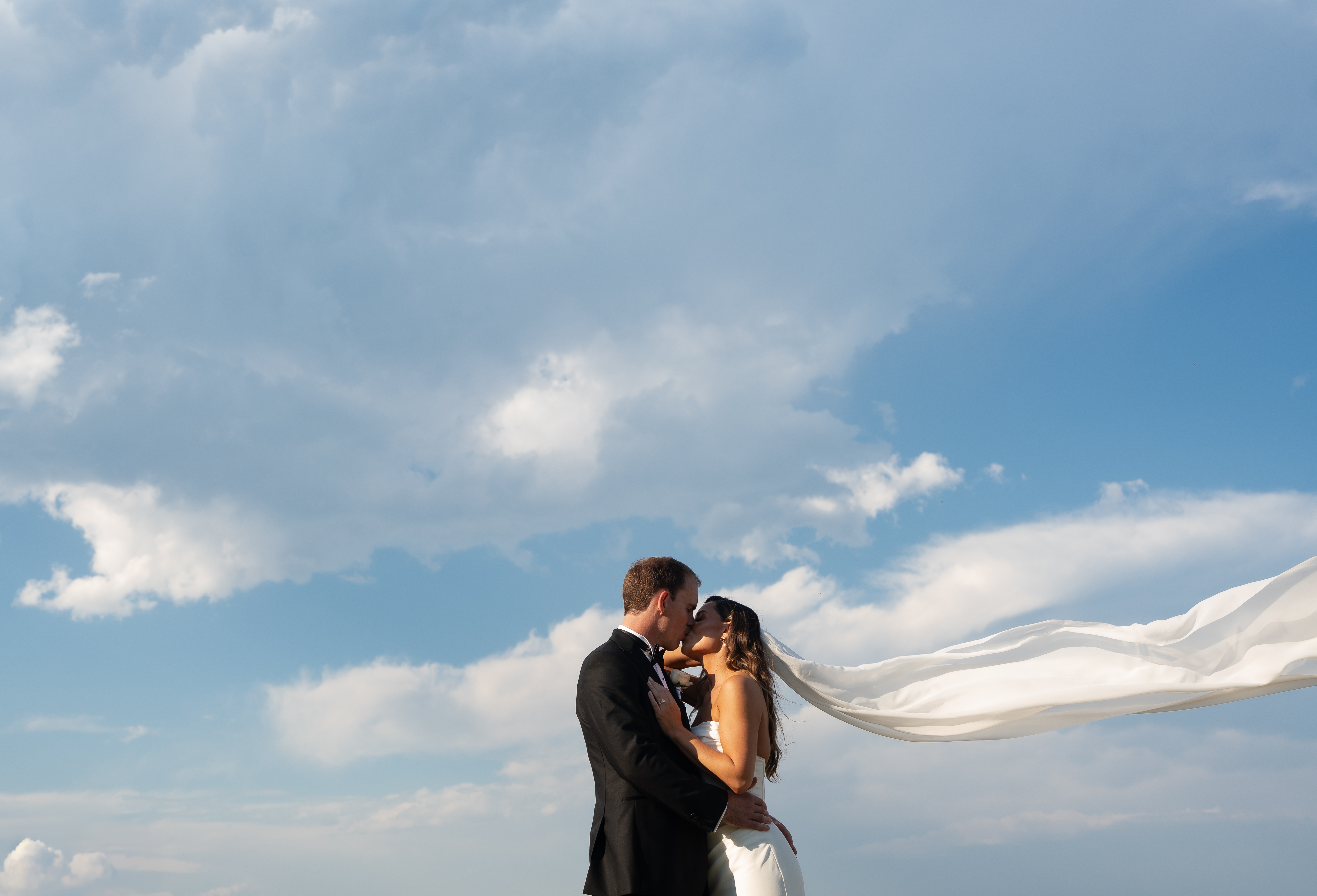 Couple on the beach, her veil pulled sideways by the wind under a big cloud sky