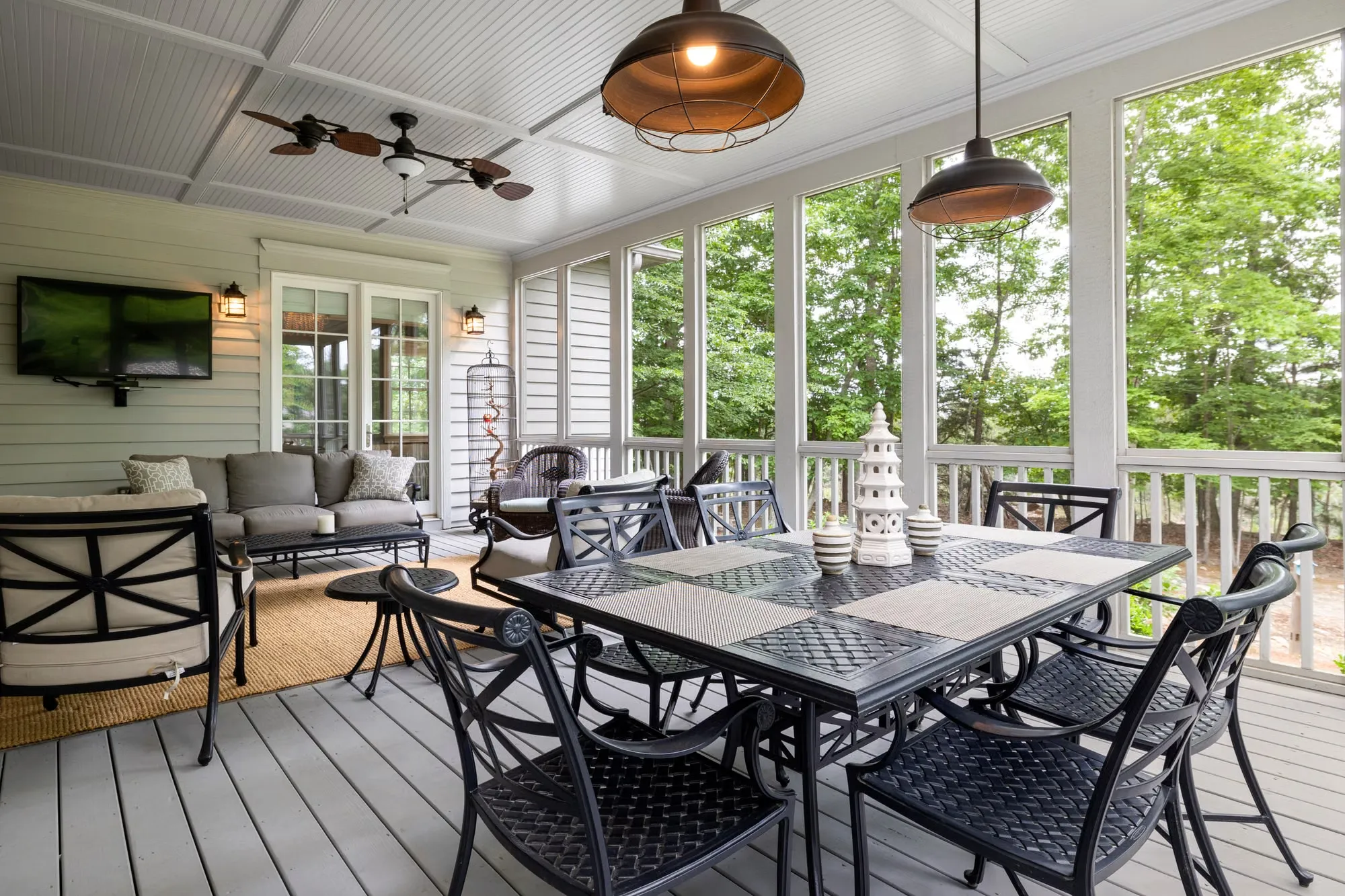 Screened dining porch with a mounted TV, sofa seating and pendant lighting overlooking a wooded backyard