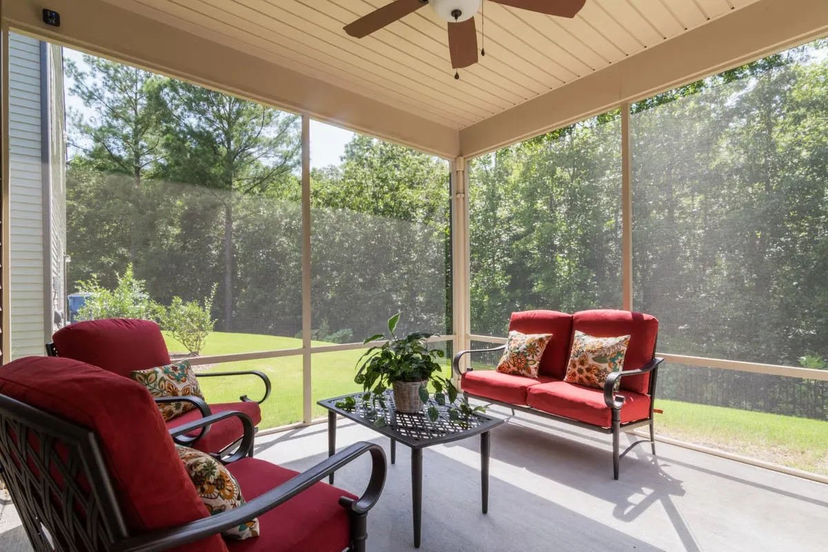 Screened porch with red cushioned furniture and a ceiling fan, overlooking a lawn and trees