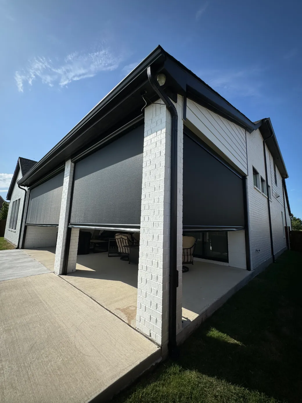 Retractable exterior shade screens lowered over a covered patio, mounted between painted brick columns under a gable roof