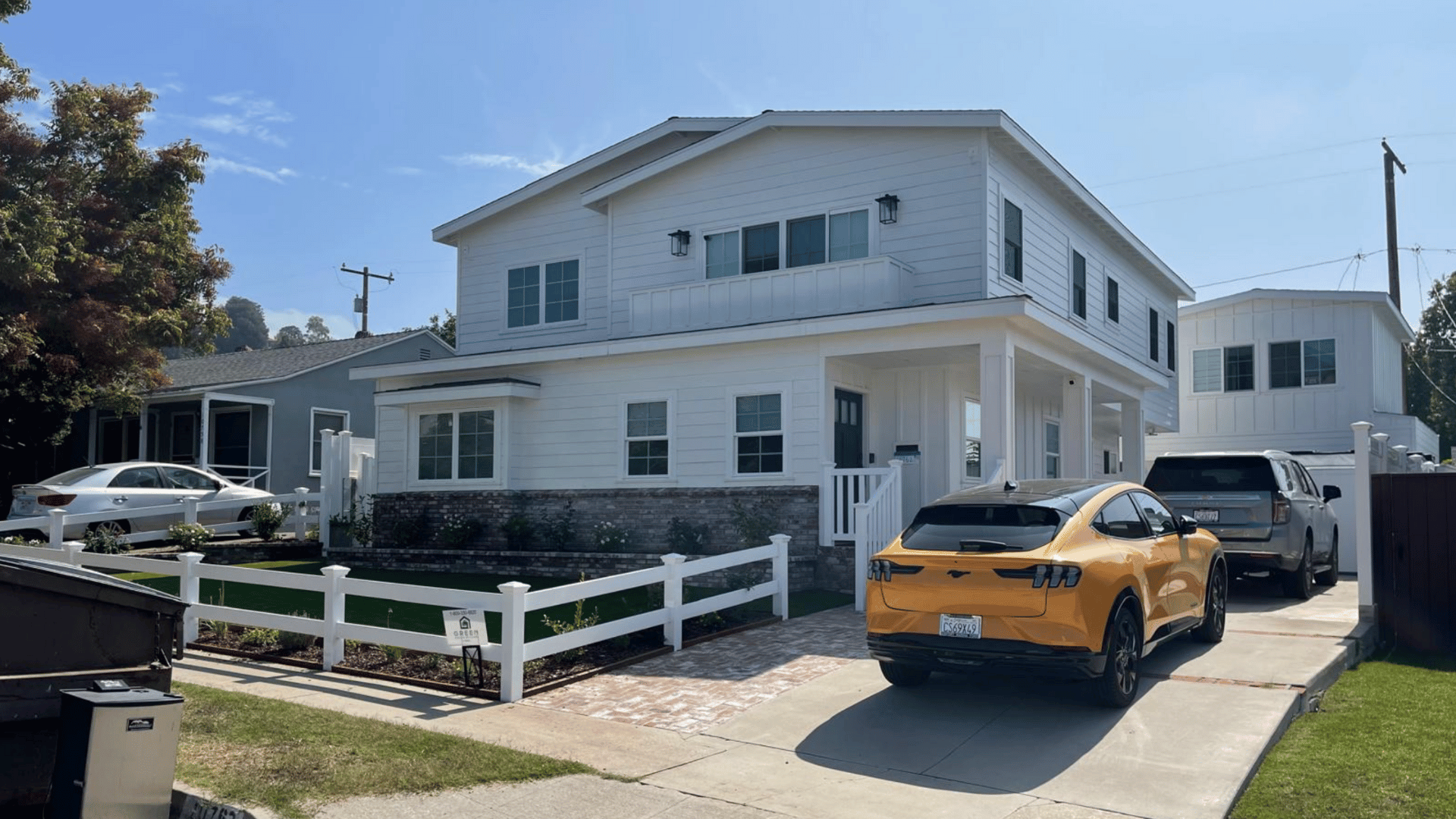 Two-story white house with a wraparound porch, brick accents, and driveway with parked vehicles.