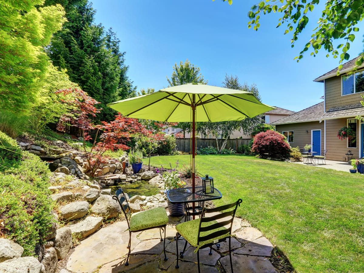 Peaceful backyard with a small seating area under a green umbrella, surrounded by lush landscaping, including a rock garden and well-maintained lawn.