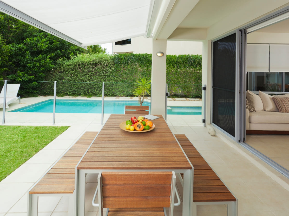 Covered patio dining area adjacent to a small, private pool, with a view of a neatly trimmed hedge and outdoor lounging space.