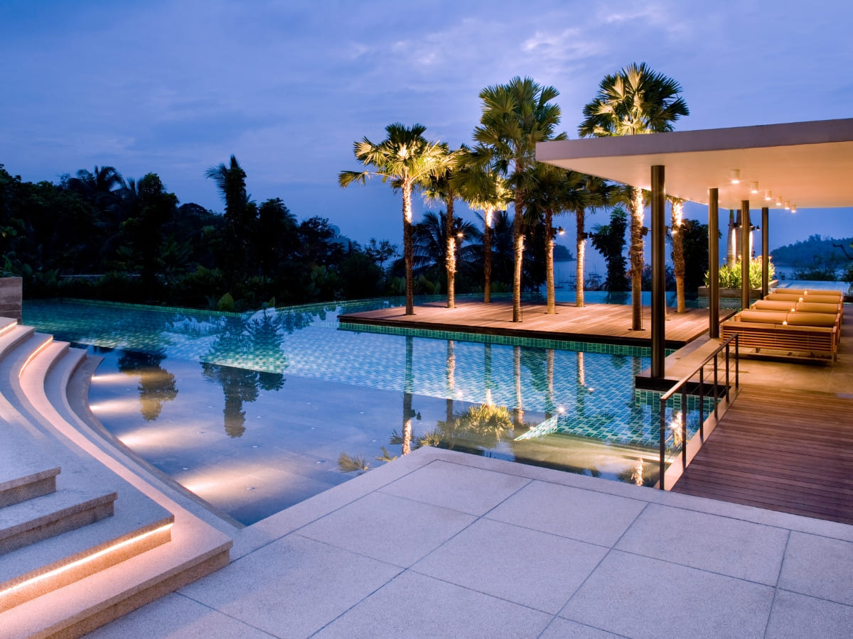 Modern infinity pool with steps, surrounded by palm trees and an outdoor deck area, beautifully lit in the evening.