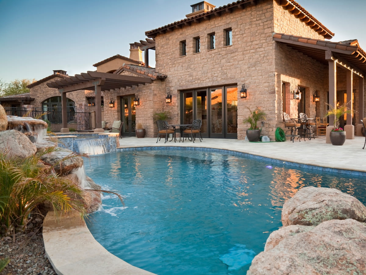 Mediterranean-style home with a large, luxurious pool featuring rock waterfalls and adjacent outdoor dining area under a wooden pergola.