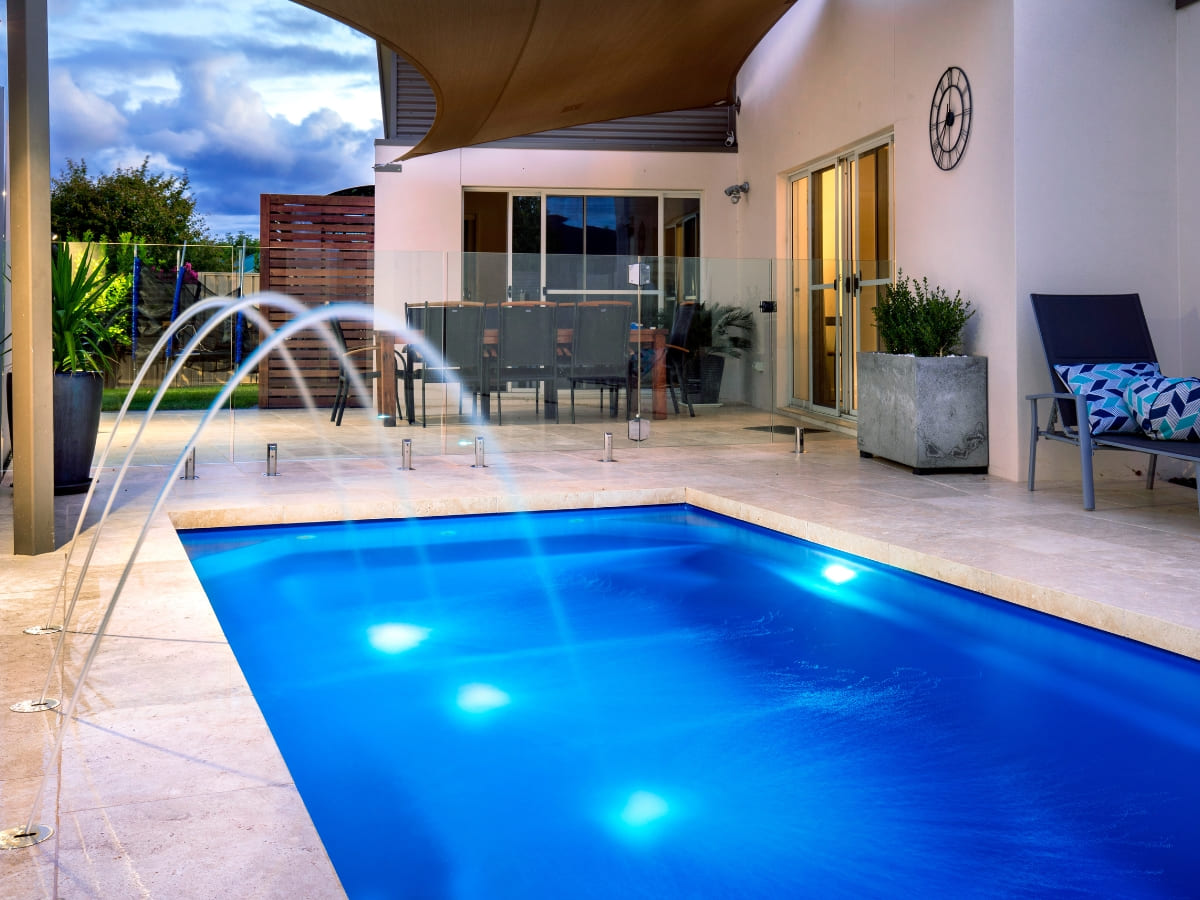 Contemporary small outdoor pool with blue lighting and water features, surrounded by a glass fence and adjacent to a patio dining area.