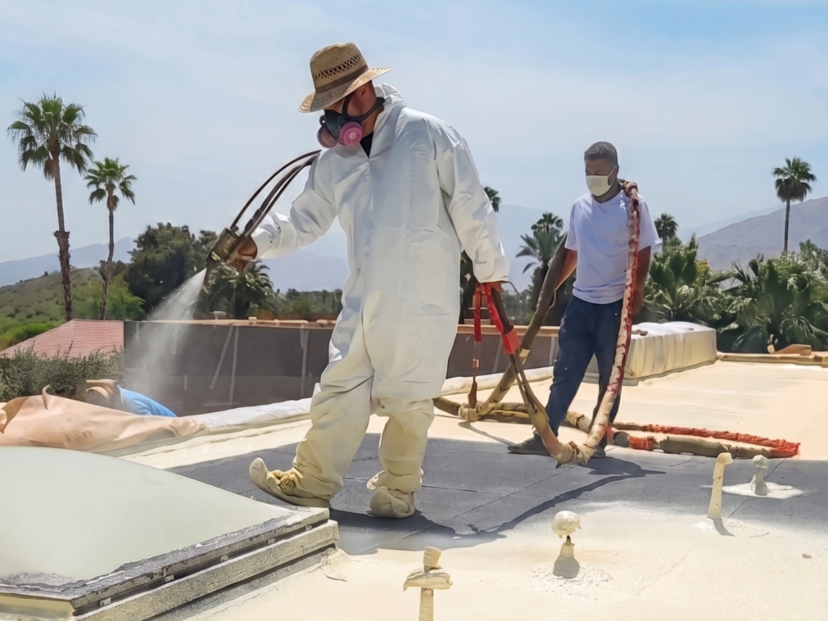 Two roofing workers in protective gear applying spray foam on a flat rooftop, with palm trees and desert mountains in the background Two roofing workers in protective gear applying spray foam on a flat rooftop, with palm trees and desert mountains in the background