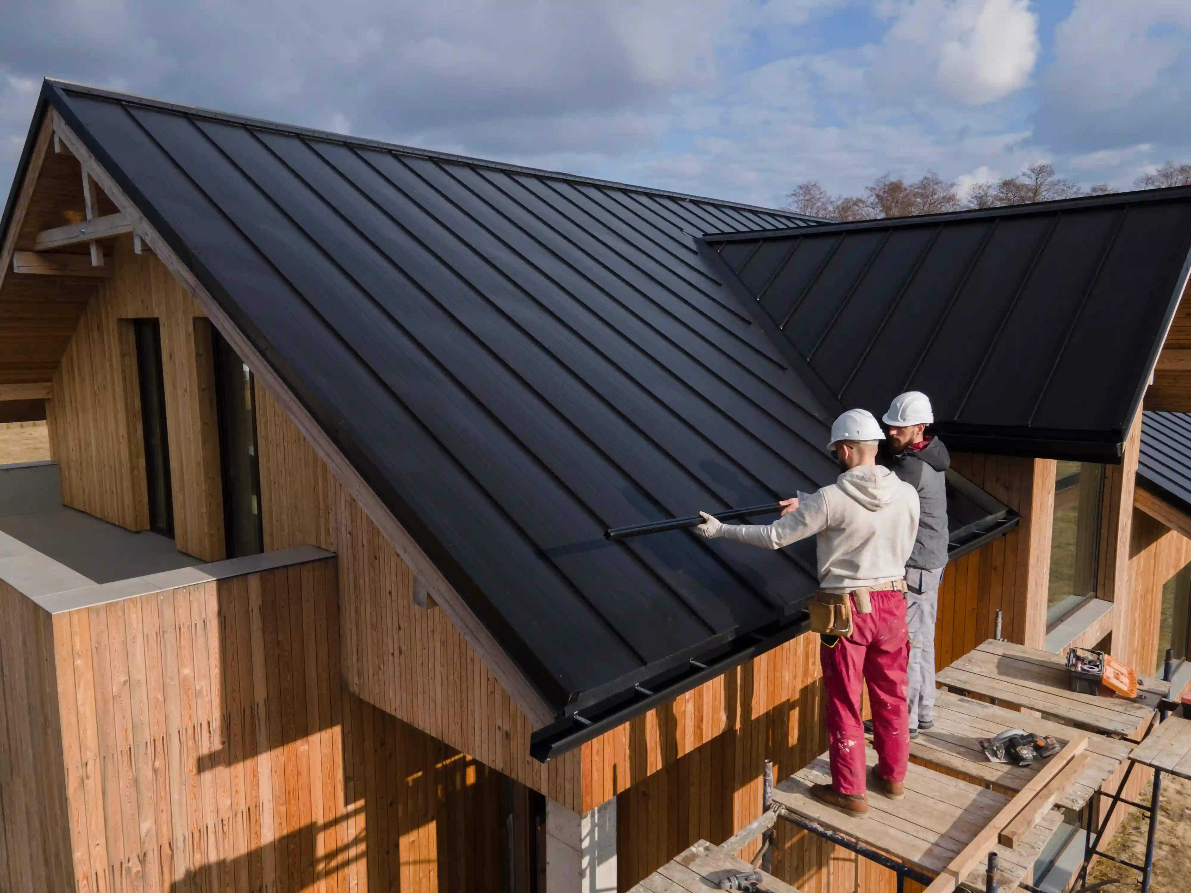 Two roofers installing black standing-seam metal roofing on a modern home
