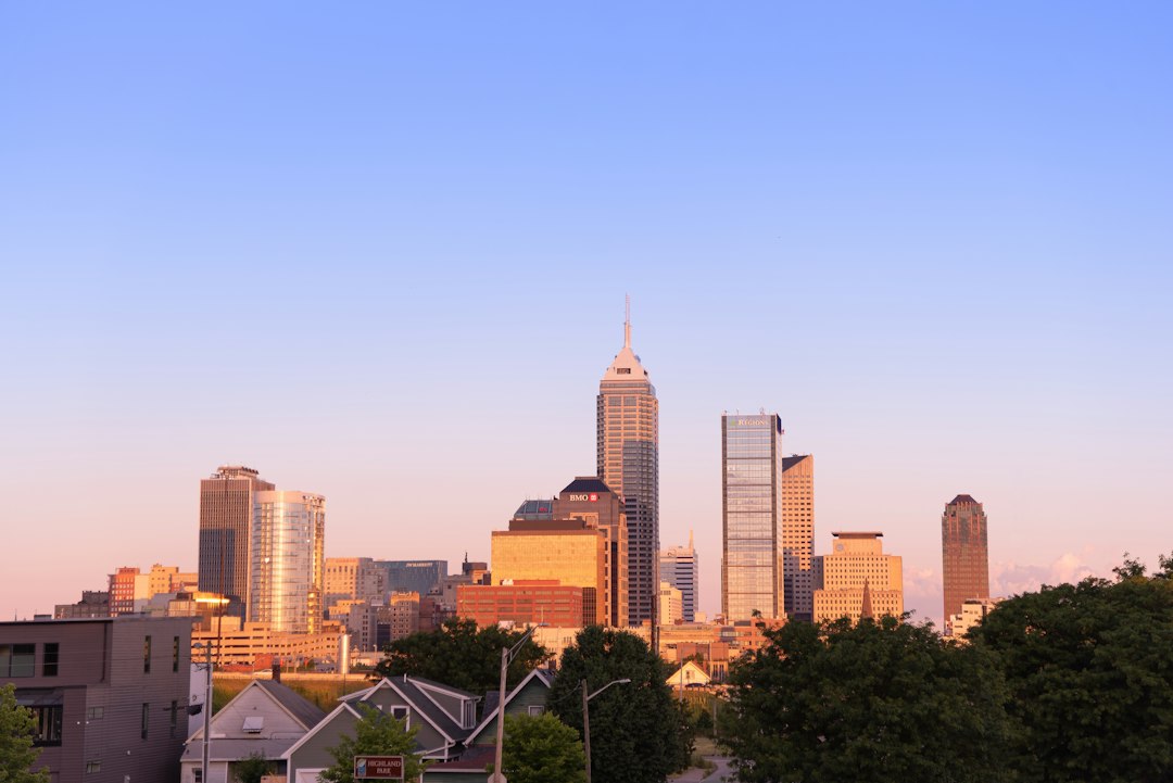 Indianapolis skyline at sunrise seen over treetops and rooftops, with a clear pale blue and pink sky.