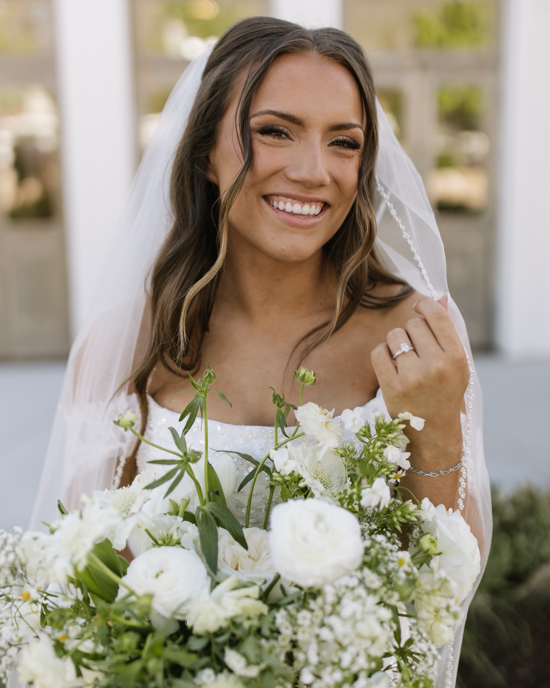 Smiling bride in a cathedral veil holding a lush white garden rose and greenery bouquet