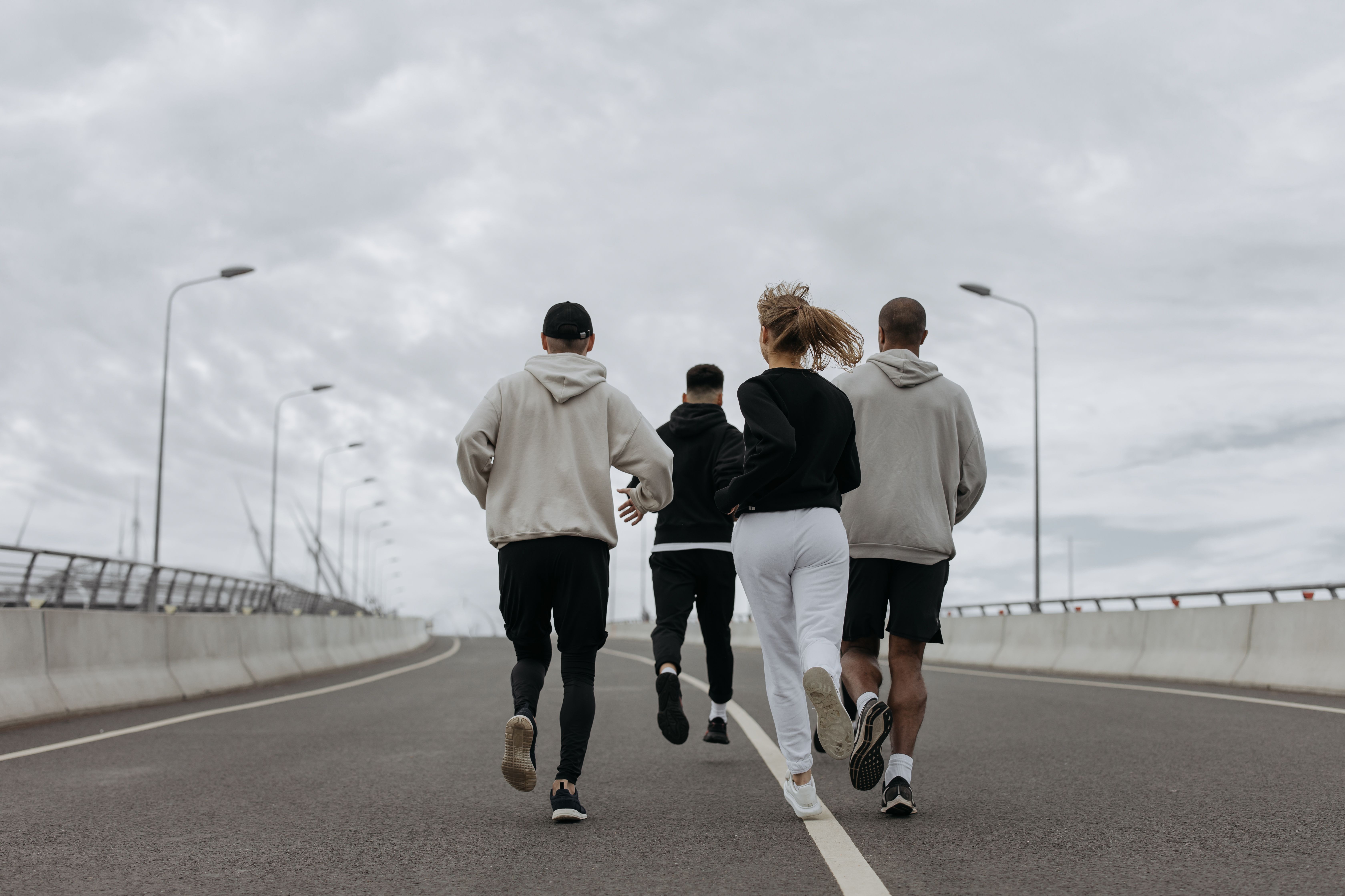 A group of four adults in athletic clothing running together on an open road under a cloudy sky, viewed from behind.