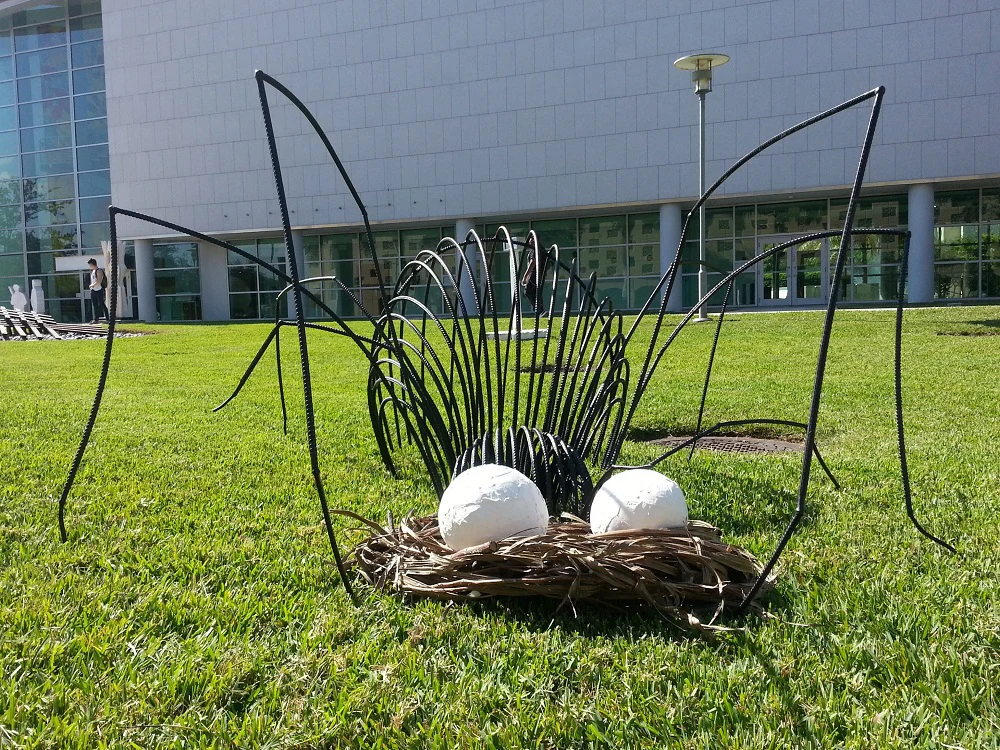 Large black metal sculpture of a spider standing over two white spherical objects on a green lawn, with a modern glass building in the background.
