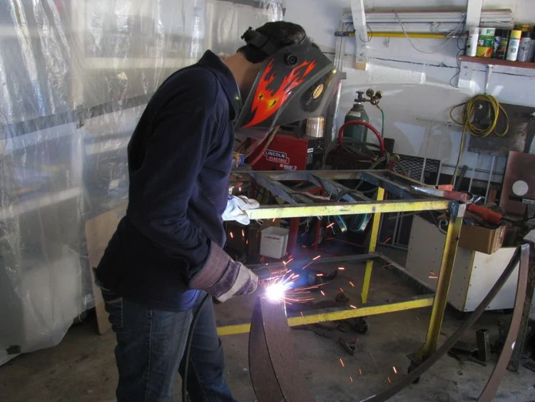 Person wearing a dark blue shirt and a red welding mask, actively welding a metal frame structure in a workshop, bright blue sparks visible.