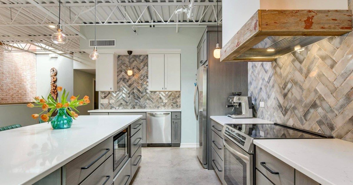 Kitchen with gray cabinets, a white island, a herringbone tile backsplash and a rustic wood range hood under an open industrial ceiling