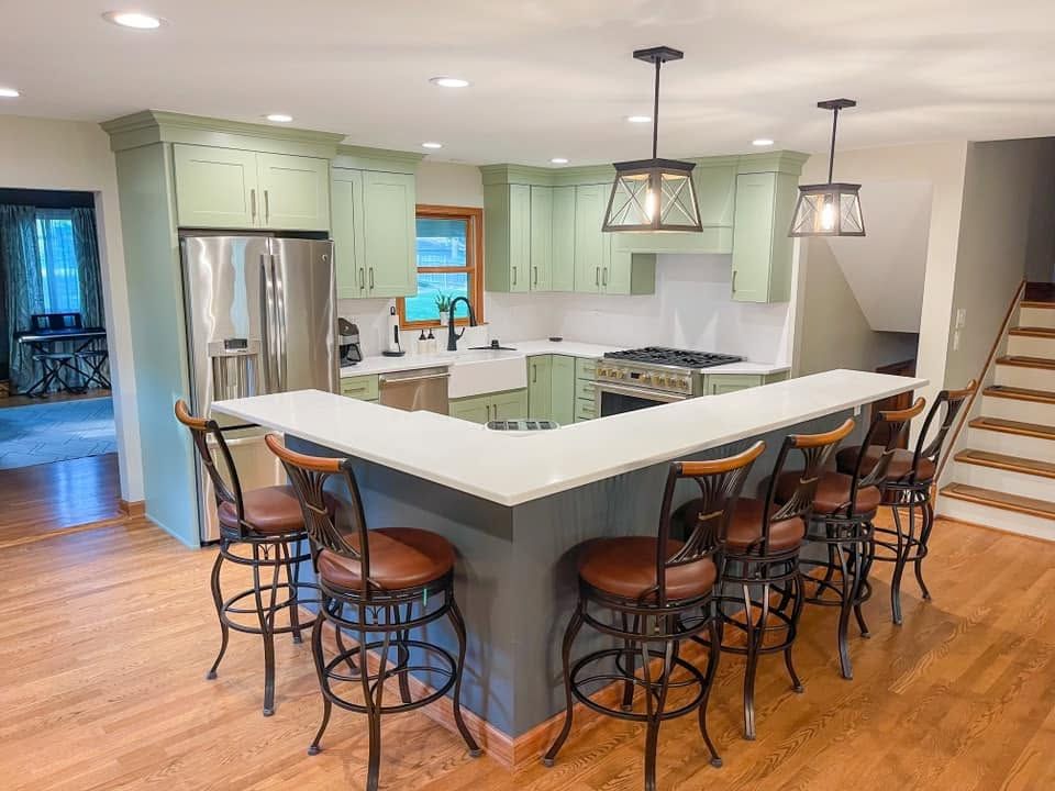 Sage green shaker kitchen with an L-shaped white counter, six leather counter stools and lantern pendant lights