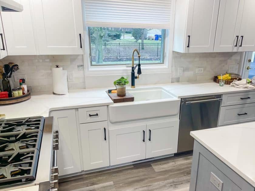 White shaker kitchen with a farmhouse sink under a wide window, a brass and black faucet and a gray island in the foreground