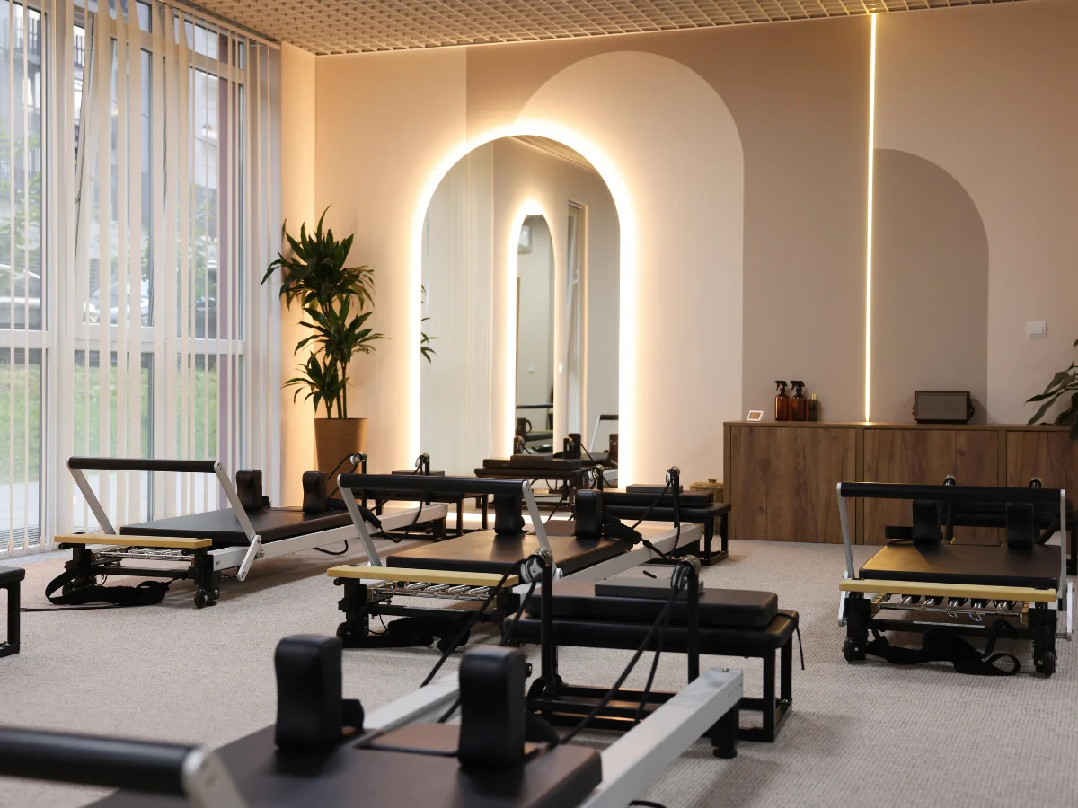 A modern, well-lit pilates studio with several black and white reformer machines on a light carpeted floor.