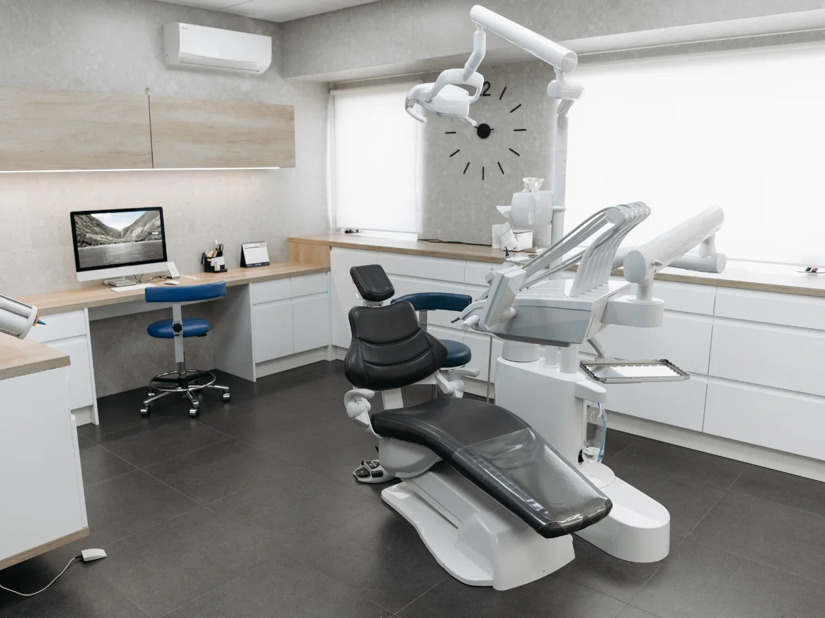 A clean, modern dental office interior with a black dental chair, equipment, and a desk with a computer.