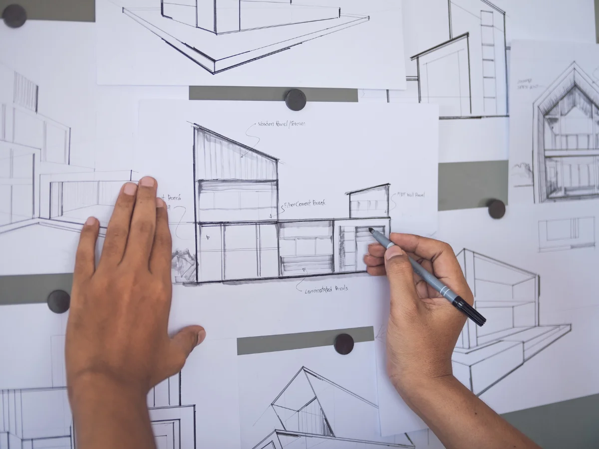Overhead view of hands sketching modern architectural house designs on papers pinned to a board.