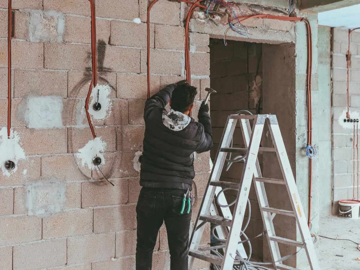 An electrician hammers an electrical conduit into a concrete block wall during construction, with a ladder nearby.