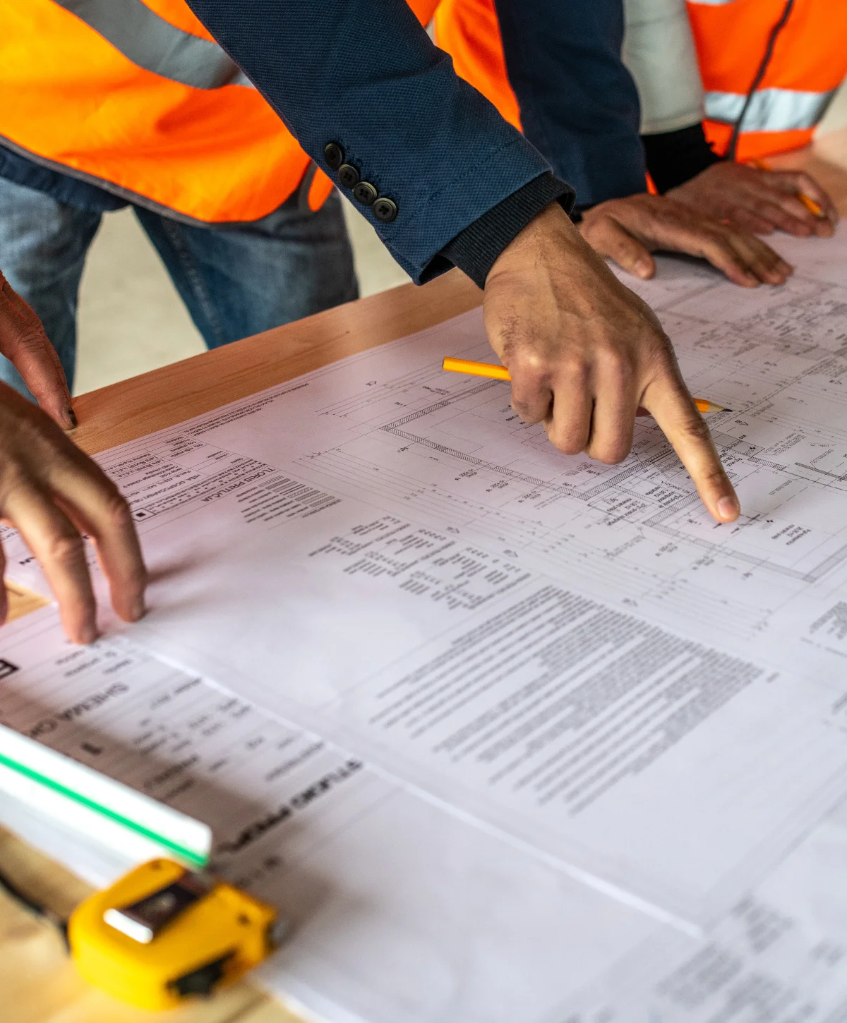 Two people in safety vests review building blueprints on a wooden table, with one person pointing at a detail.