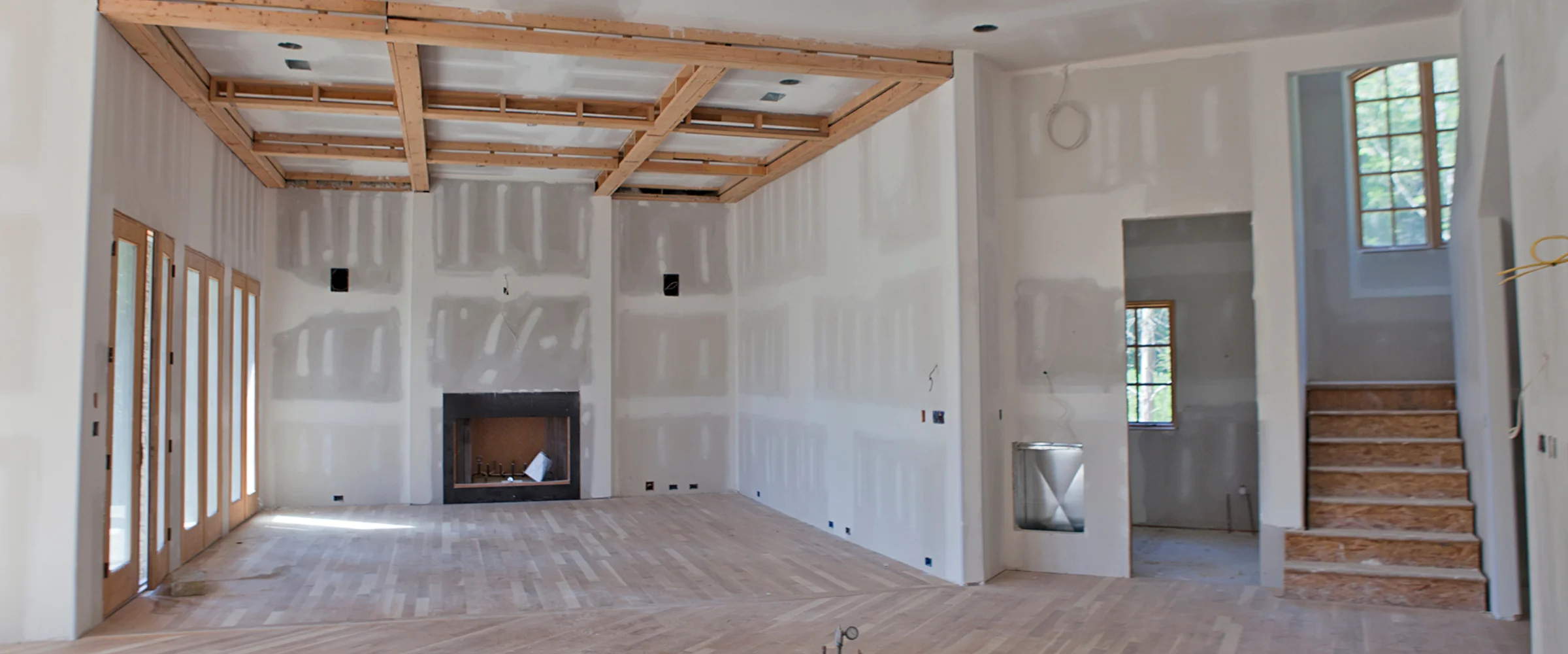 An unfinished home interior showing drywall, exposed ceiling beams, hardwood floor, fireplace, and staircase.