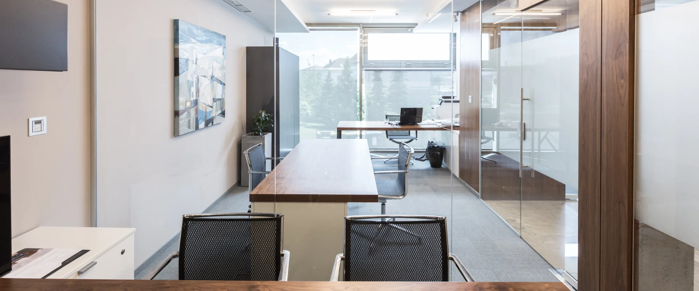 A modern office interior featuring a long wooden table, mesh chairs, and a glass partition separating two workspaces.
