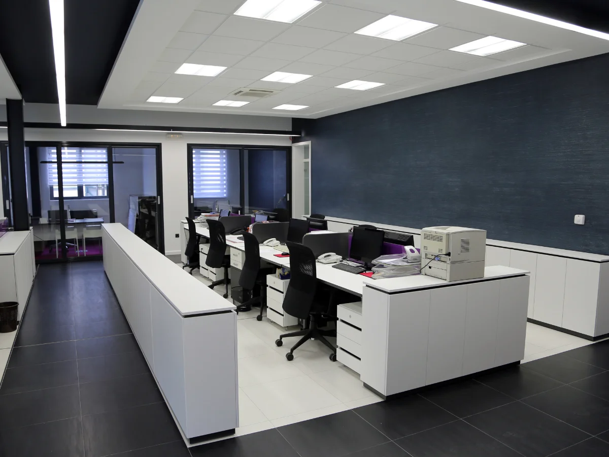 A modern open-plan office with white desks, black chairs, and computers against a dark blue textured wall.