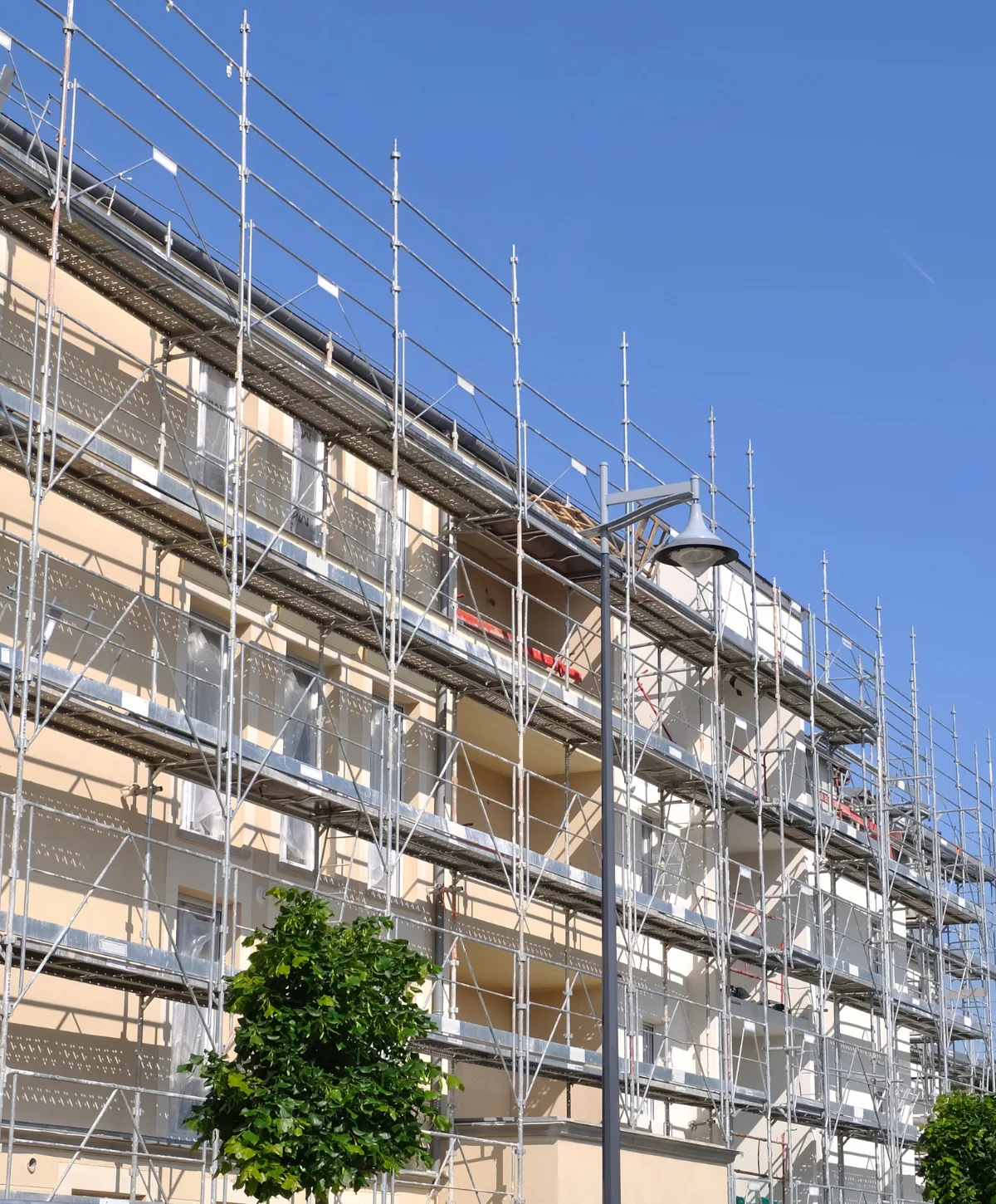 A beige building facade covered in extensive metal scaffolding under a clear blue sky, with a street light and green trees.