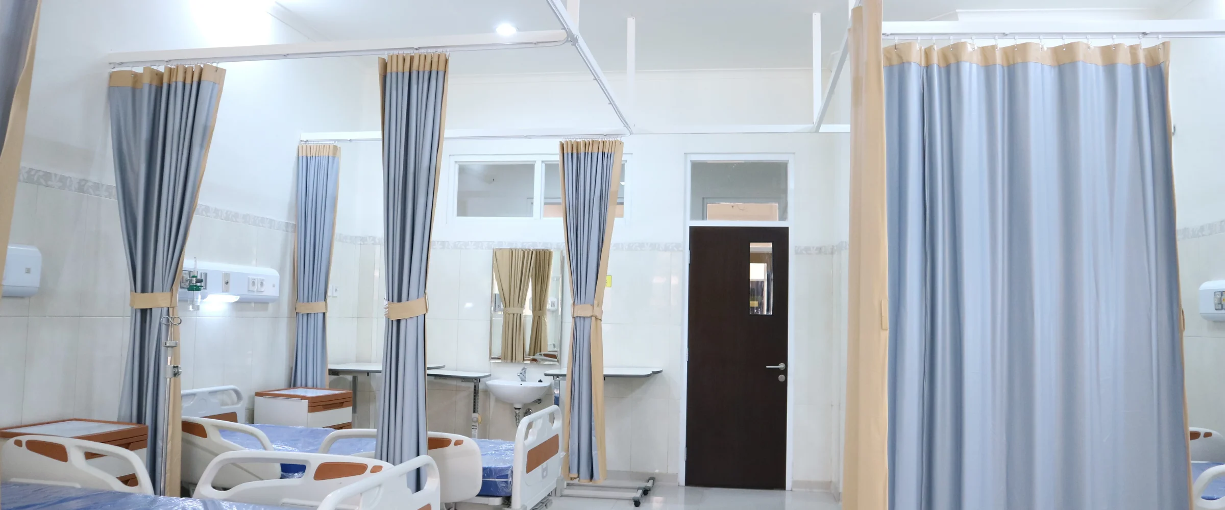 A clean, modern hospital room with multiple beds, blue privacy curtains, a sink, and a dark brown door.