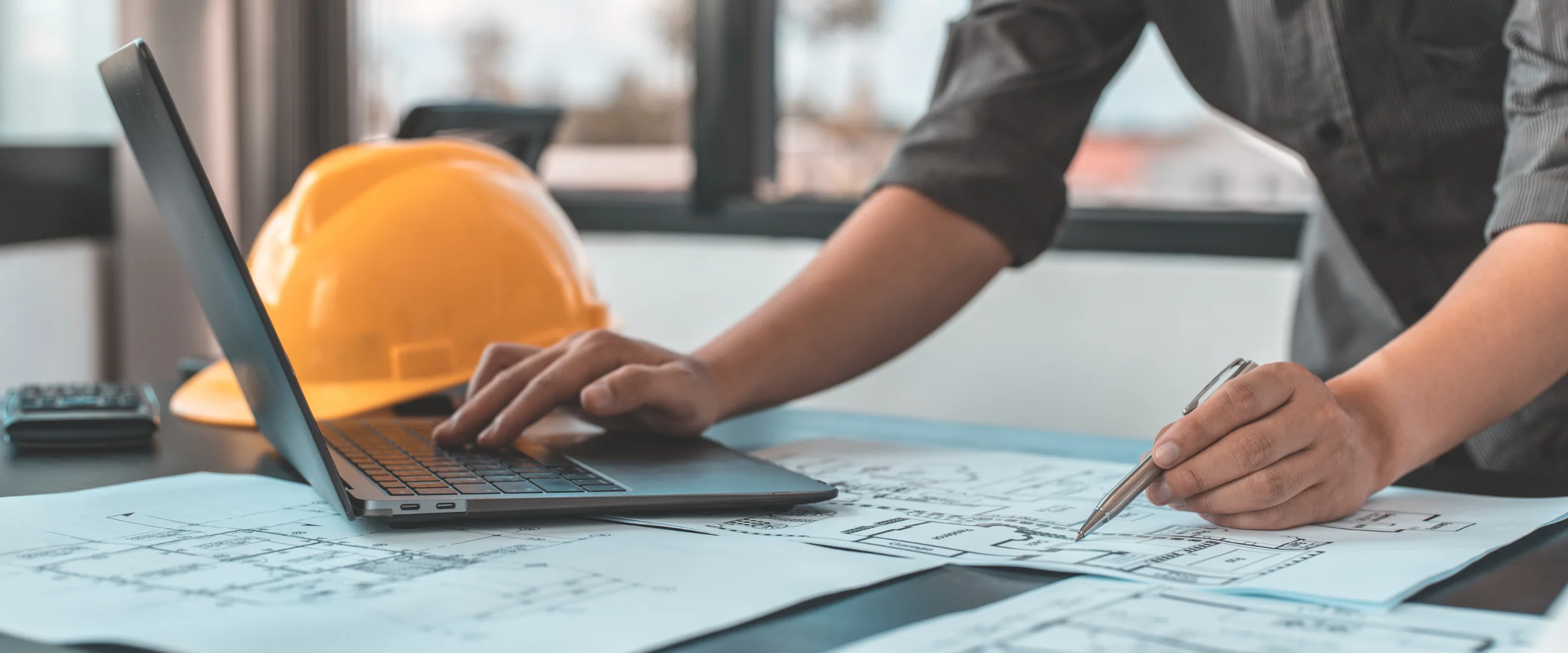 An architect types on a laptop while pointing at blueprints with a pen, with a yellow hard hat nearby.