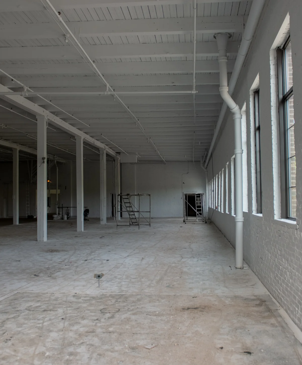 A large, empty industrial loft space with white painted brick walls, exposed ceiling beams, and concrete floors.