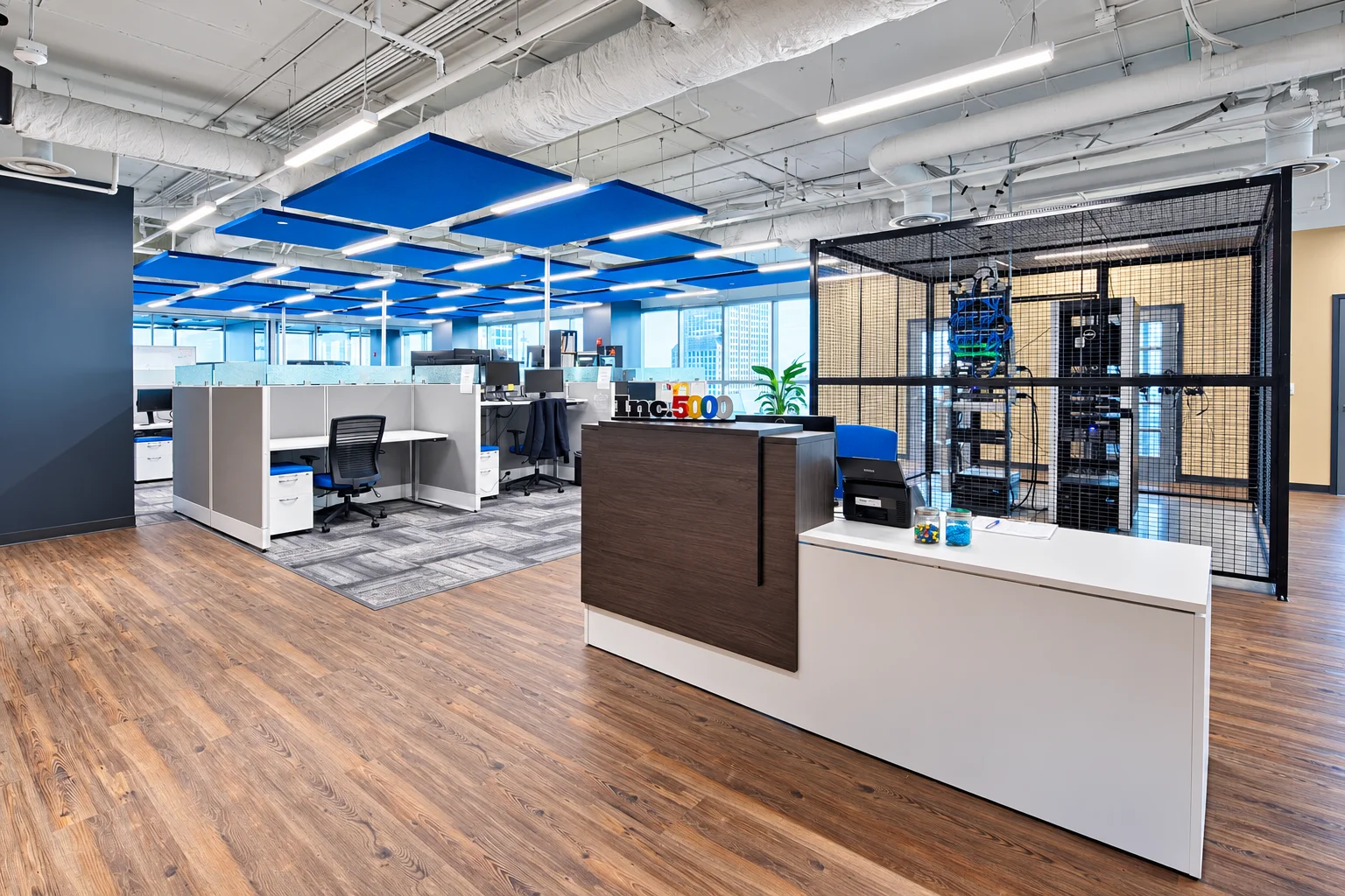 A modern open-plan office features a reception desk with an 'Inc. 5000' sign, cubicles, blue ceiling panels, and wood-look flooring.