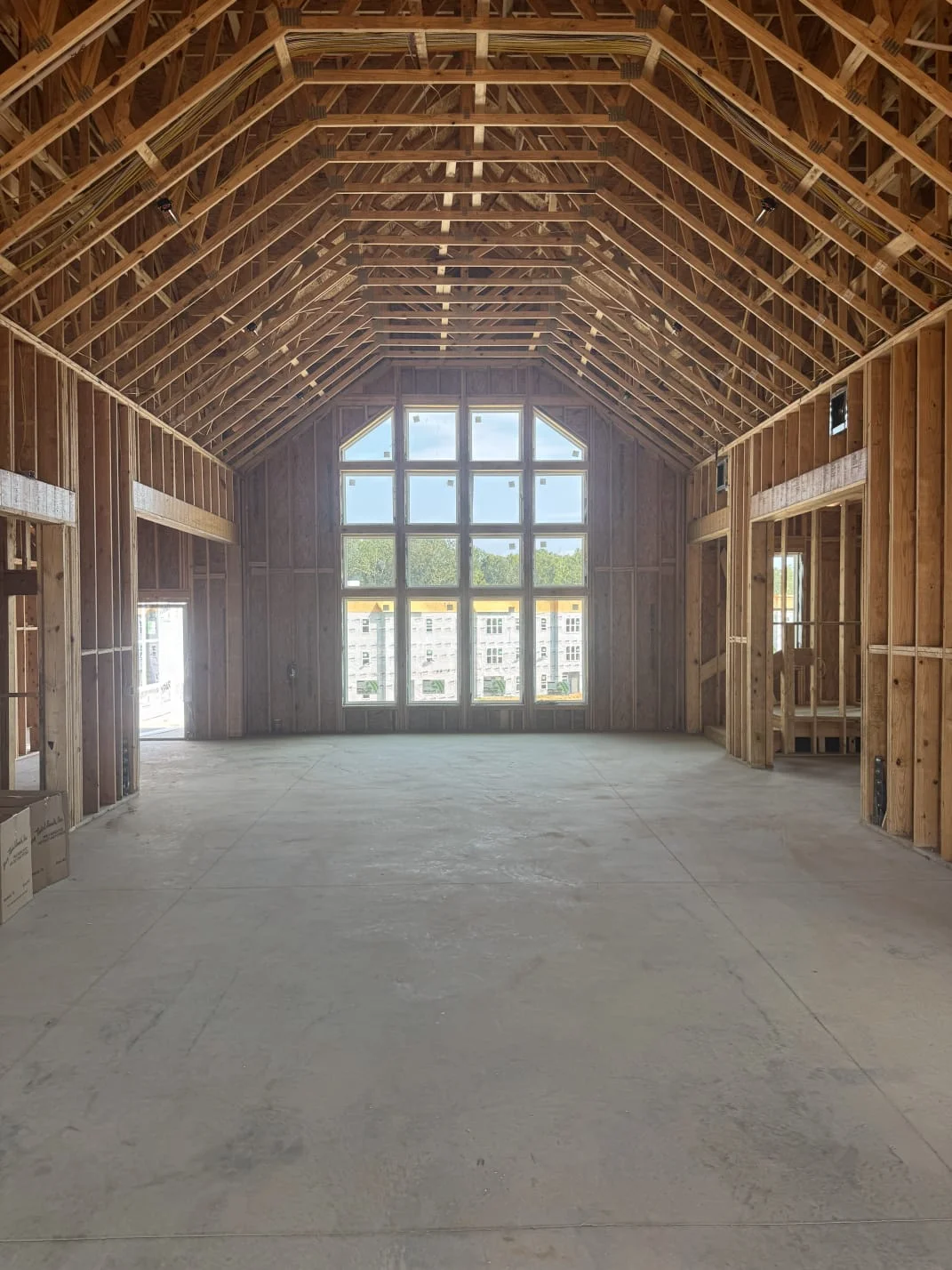 An interior view of a house under construction, showing wooden wall studs, ceiling trusses, and a large window at the far end.