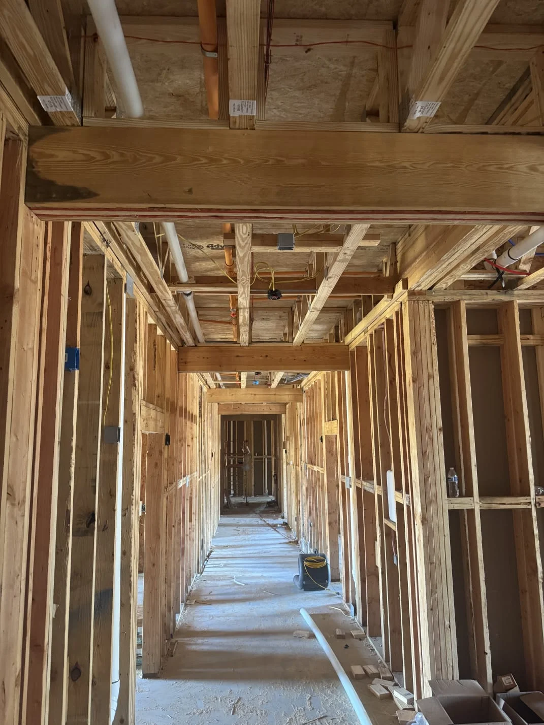 A long interior hallway of a house under construction, framed with exposed wooden studs, beams, pipes, and wiring.