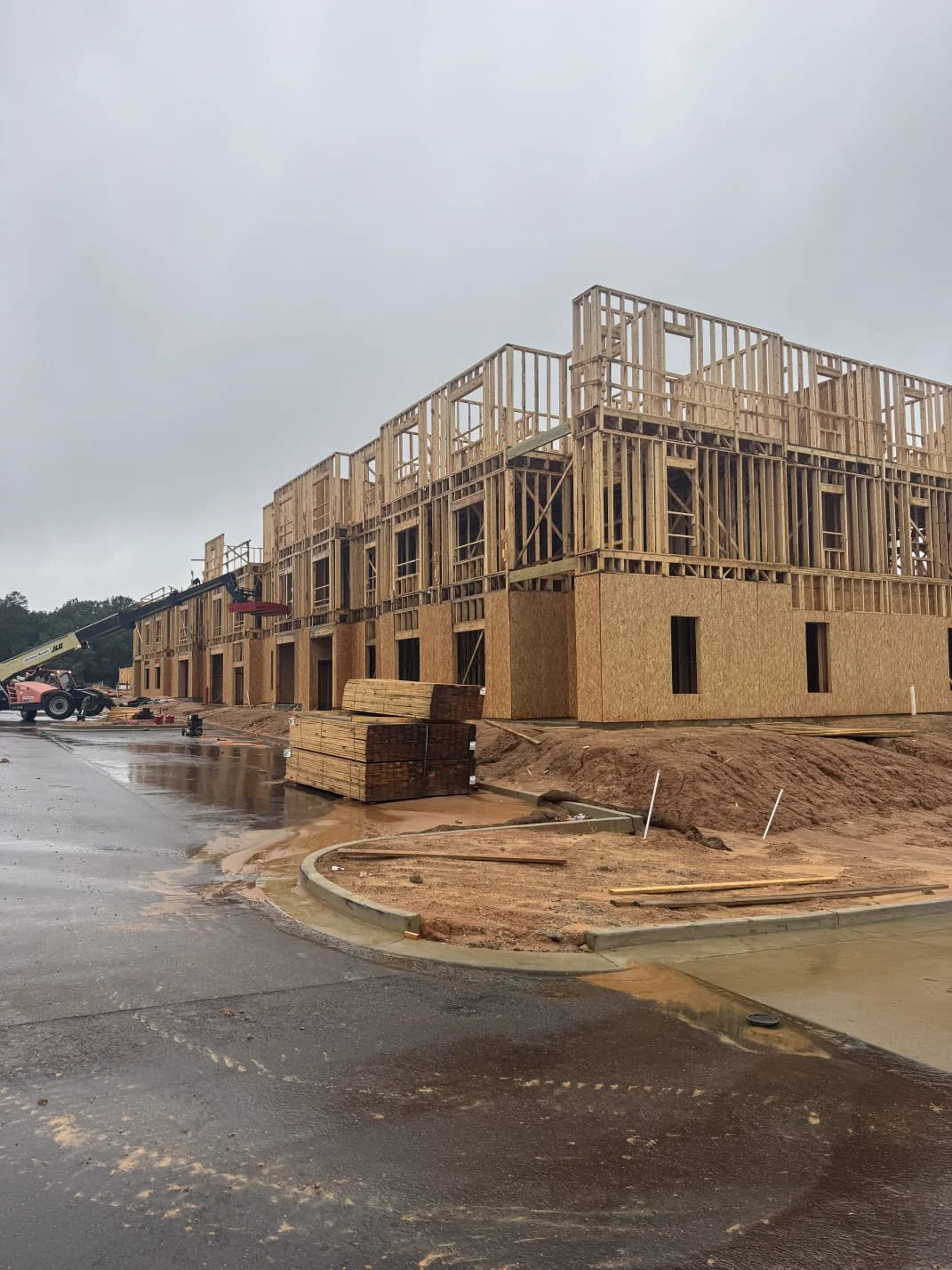 A multi-story building under construction with wood framing and plywood sheathing on a wet, cloudy day.