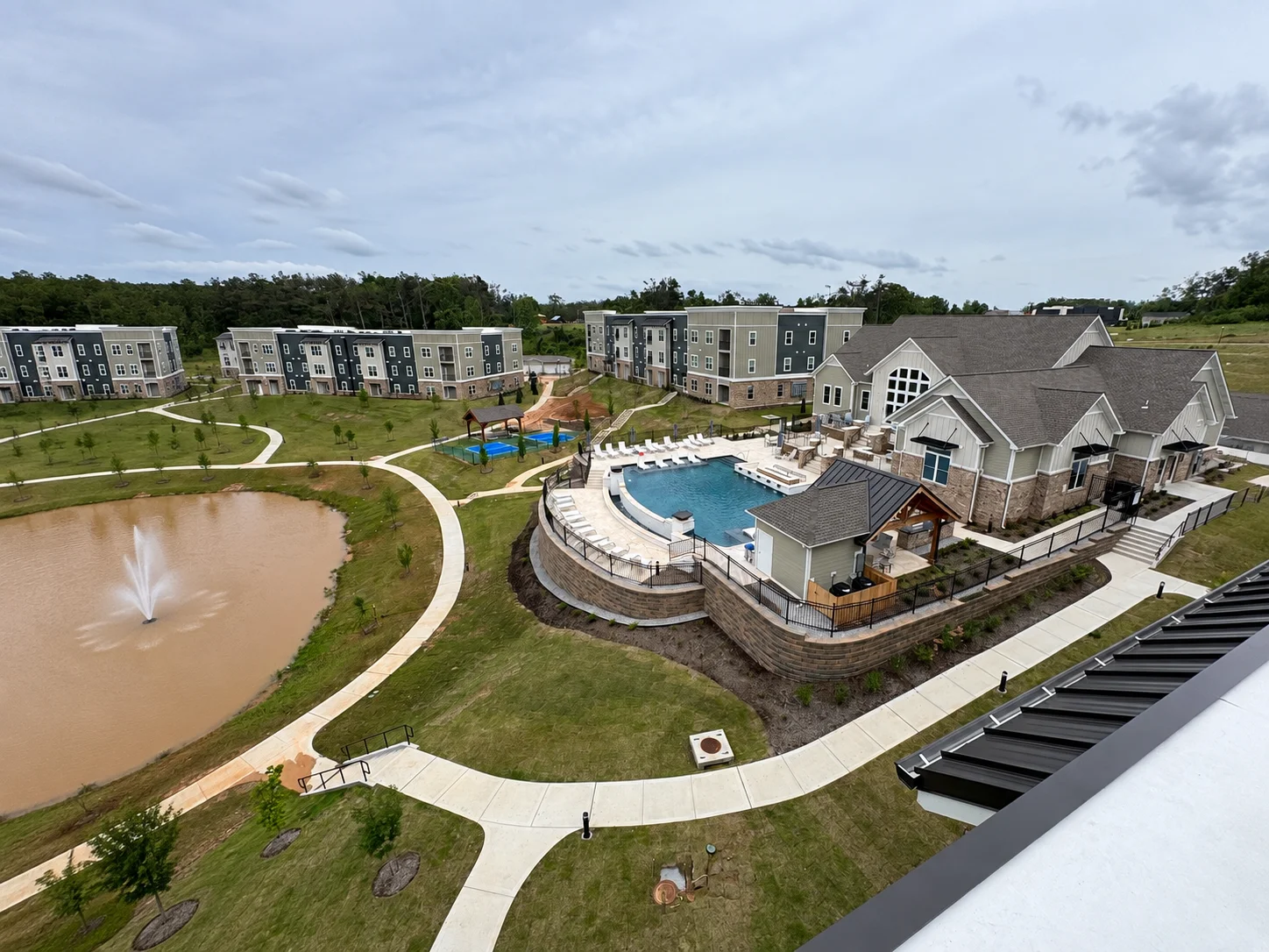 An aerial view of a modern apartment complex featuring a large pool, clubhouse, and winding path next to a pond under a blue sky.