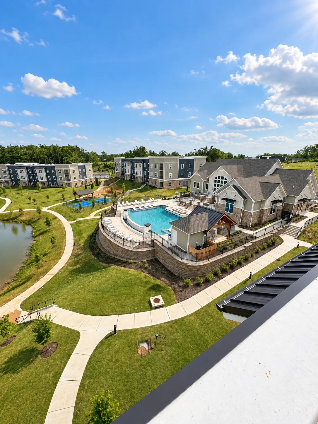 An aerial view of a modern apartment complex featuring a large pool, clubhouse, and winding path next to a pond under a blue sky.