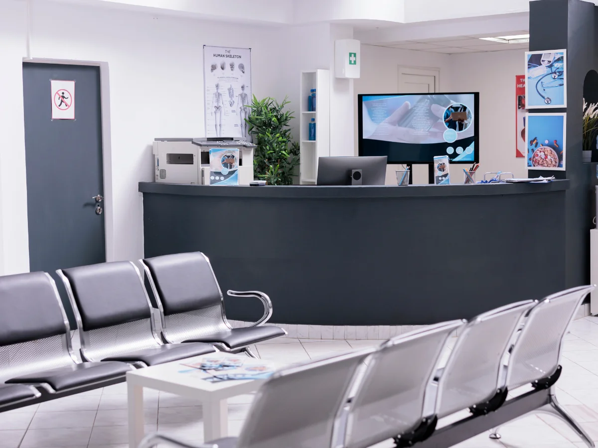 An empty, modern medical clinic waiting room with a dark grey reception desk, multiple chairs, and a screen displaying medical information.