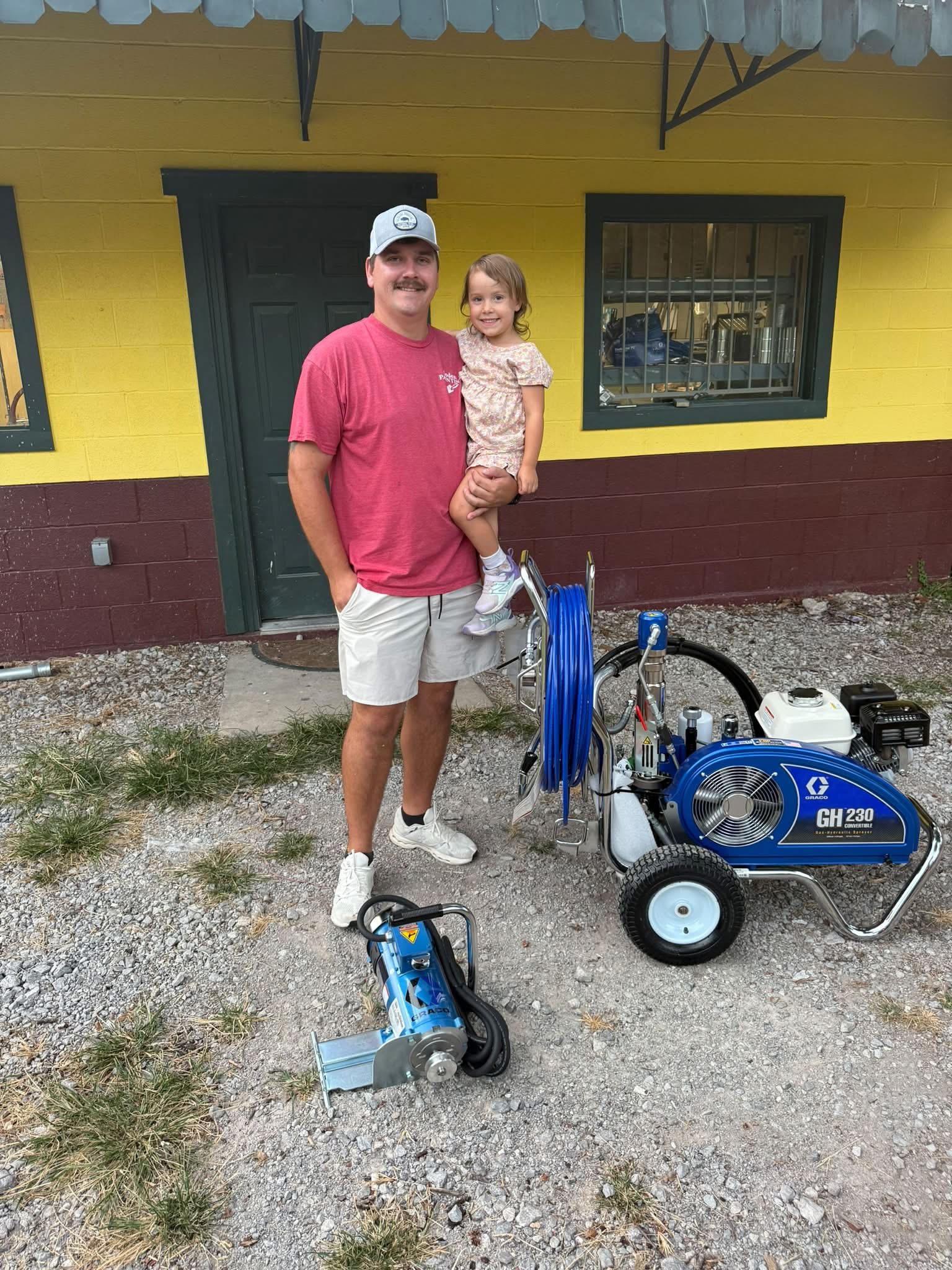 A man holding a child outside the storefront, beside painting equipment.